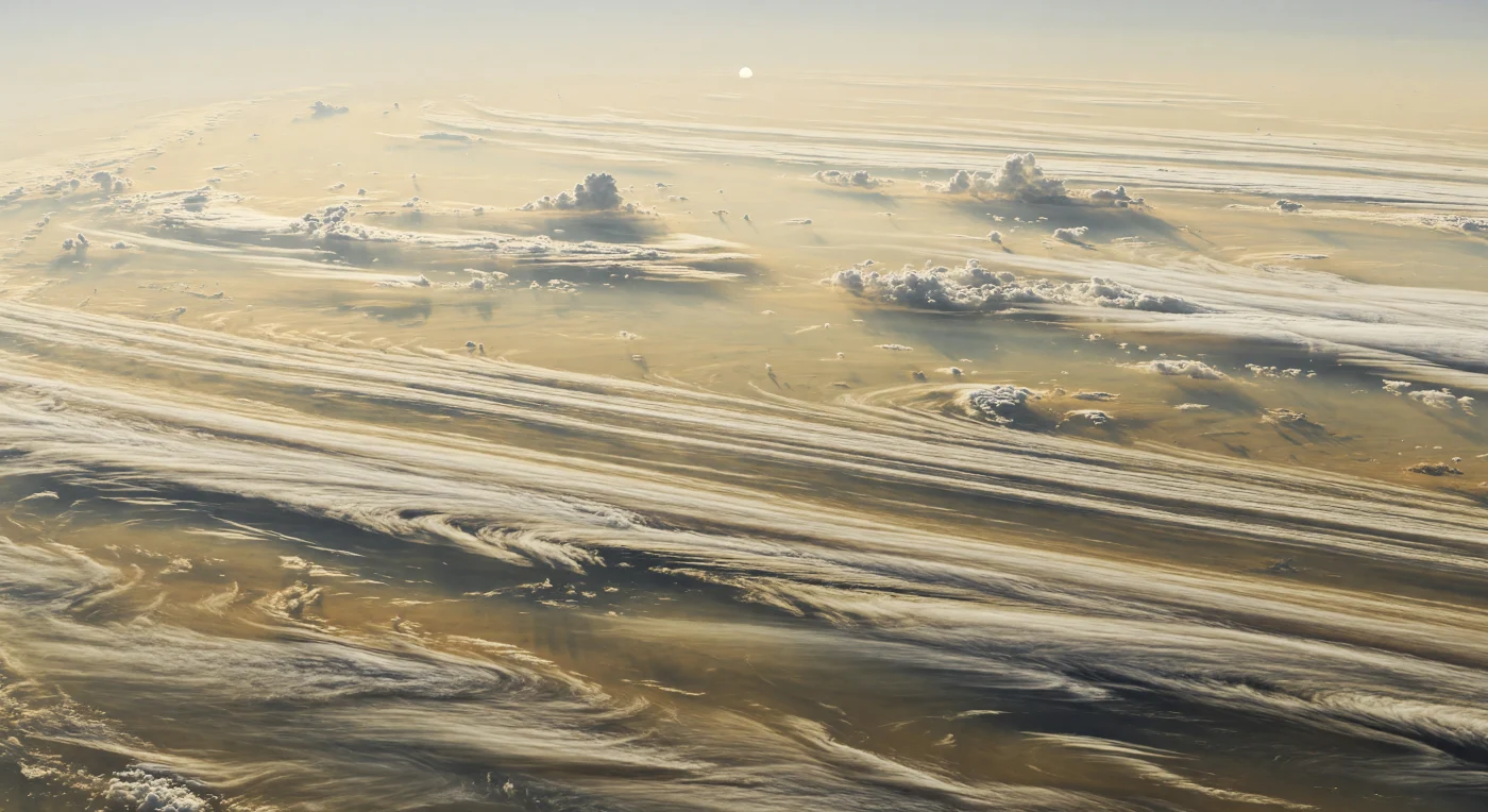من هذا الموضع العائم قرب مستوى الضغط القريب من 1 بار، لا تظهر أرض على الإطلاق، بل سهول هائلة من السحب تمتد حتى الأفق على هيئة أشرطة متوازية من العسلي الفاتح، والعاجي، والبيج، والذهبي الرمادي، وقد مشّطتها رياح نفاثة شديدة السرعة إلى خيوط ريشية وتموجات مضفّرة ودوامات بيضوية خافتة. بين هذه الأشرطة المضيئة تنفتح ممرات أغمق بلون البرونز الرمادي والمغرة الدخانية، تمثل طبقات سحابية أعمق يُرجّح ارتباطها بمواد مثل هيدروسلفيد الأمونيوم، بينما تتكوّن القمم الأعلى من بلورات جليد الأمونيا المعلّقة في غلاف غازي بلا سطح صلب ولا جبال ولا بحار. الضوء هنا خافت ودافئ على نحو مخادع، إذ يتسرّب قرص الشمس الصغير والبعيد عبر ضباب هيدروكربوني وضباب ضوئي كيميائي يلطّف التباين ويغمر المشهد بتوهج كريمي باهت، فيما تبدو الخلايا العاصفة البعيدة صغيرة على نحو مدهش أمام أحزمة جوية بحجم قارات. ومع انحناء الأشرطة السحابية تدريجيًا في البعيد واختفائها داخل العتمة الضبابية، يصبح الإحساس بالمقياس كاسحًا: أنت لا تنظر إلى طقس فوق عالم، بل إلى عالم مؤلف بالكامل تقريبًا من الطقس.