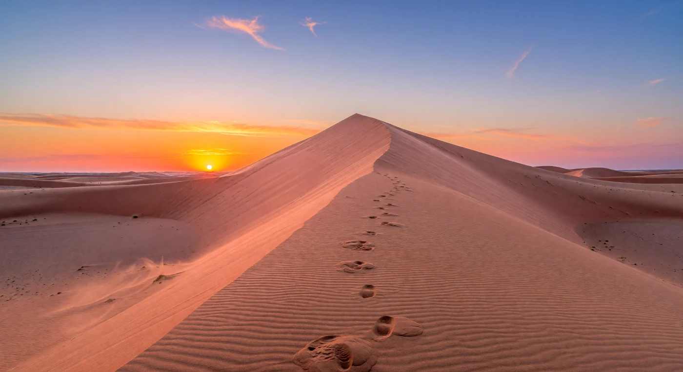 Am Fuß einer gewaltigen Erg-Düne blickt man eine rund 200 Meter hohe Wand aus feinkörnigem, gut sortiertem Quarzsand hinauf, deren messerscharfer Kamm sich dunkel gegen den glühenden Abendhimmel abzeichnet. Das tiefe Sonnenlicht lässt die Luvseite in einem physikalisch plausiblen Farbverlauf aufleuchten – von rosigen und pfirsichfarbenen Partien nahe dem Grat über Apricot und sattes Orange bis zu rotvioletten Schatten am Dünenfuß –, während Rippelstrukturen, kleine Lawinenspuren und einzelne Fußabdrücke die ständige Arbeit des Windes sichtbar machen. Am fernen Horizont steht die etwa ,53 Grad große Sonne, durch Brechung in der dichten Atmosphäre leicht abgeflacht, und taucht die staubige Luft in Bänder aus Orange, Magenta und Violett, die nach oben in ein klares, tiefes Blau übergehen. In dieser trockenen, vegetationslosen Weite aus mineralischem Sand, warmem Dunst und langen, scharf gezeichneten Schatten wird die enorme Maßstäblichkeit der Dünenlandschaft unmittelbar spürbar, als stünde man selbst im stillen Atem eines riesigen Wüstenmeeres.
