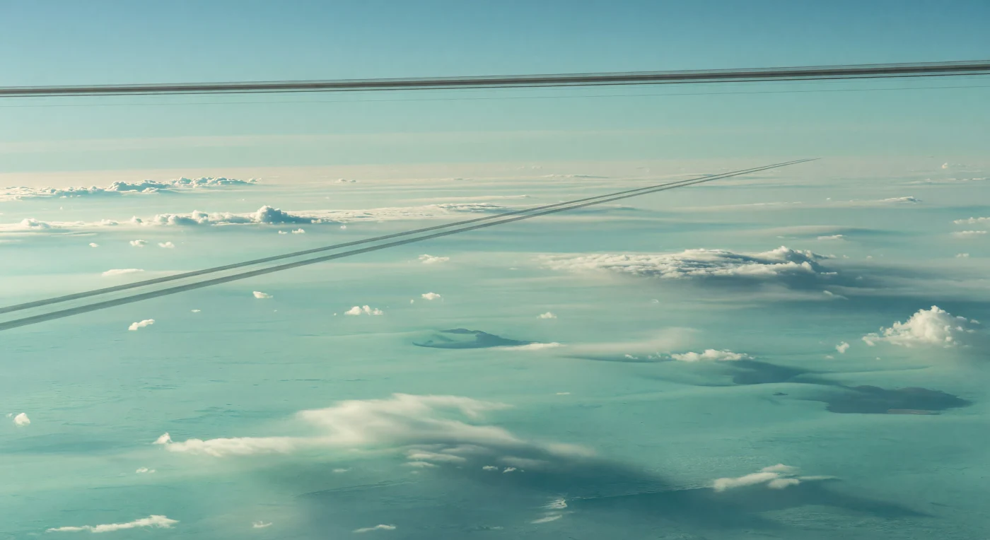Depuis les hautes couches de l’atmosphère, on flotte au-dessus d’une plaine nuageuse infinie d’un aigue-marine laiteux, modelée en nappes douces, brumes stratifiées et faibles bourgeonnements convectifs, sans aucun sol ni relief solide à l’horizon. À l’équinoxe, les anneaux étroits et sombres projettent sur ce vaste pont nuageux de fines lignes d’ombre grises, parfaitement droites et parallèles, qui filent jusqu’à se perdre dans un voile turquoise, tandis qu’au-dessus de vous les anneaux eux-mêmes ne sont plus que de minces arcs anthracite suspendus dans un ciel cyan pâle. Cette géométrie tranchante contraste avec la texture souple des brumes chargées d’aérosols de méthane et d’hydrocarbures, révélant la nature d’une géante de glace où la “surface” visible n’est qu’un sommet atmosphérique autour du niveau d’environ 1 bar. Sous l’éclat minuscule et froid d’un Soleil lointain, la lumière reste diffuse, bleu-vert et faible, donnant à l’immensité courbe des nuages une présence silencieuse et irréelle, comme si l’on contemplait un océan céleste traversé par l’ombre d’une horloge cosmique.