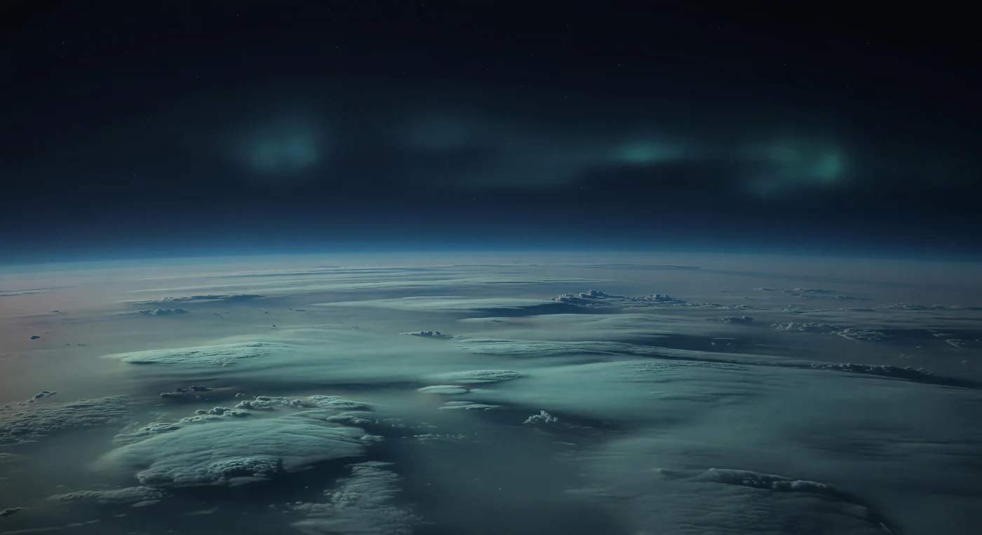 Davanti a te non c’è una superficie solida, ma un immenso mare atmosferico sospeso: banchi di nubi scure e appiattite, solcati da creste soffici, avvallamenti poco profondi e fasce troposferiche blu-nerastre che si perdono oltre un orizzonte curvo e sterminato. La foschia ricca di metano e aerosol di idrocarburi assorbe gran parte della luce, tingendo tutto di ciano pallido, turchese spento e blu d’ardesia, mentre il cielo della notte polare resta quasi nero, con le stelle appena soffocate dalla bruma alta. In alto, solo un’aurora tenue e diffusa compare come archi irregolari verdazzurri, sfumati e lattiginosi invece che brillanti tende luminose, un effetto coerente con un campo magnetico fortemente inclinato e con un’atmosfera freddissima, densa e stratificata. L’assenza di terreno, il silenzio visivo delle nubi e la profondità di questi strati che si estendono per migliaia di chilometri danno la sensazione di galleggiare dentro un crepuscolo alieno, vasto e gelido oltre ogni esperienza terrestre.