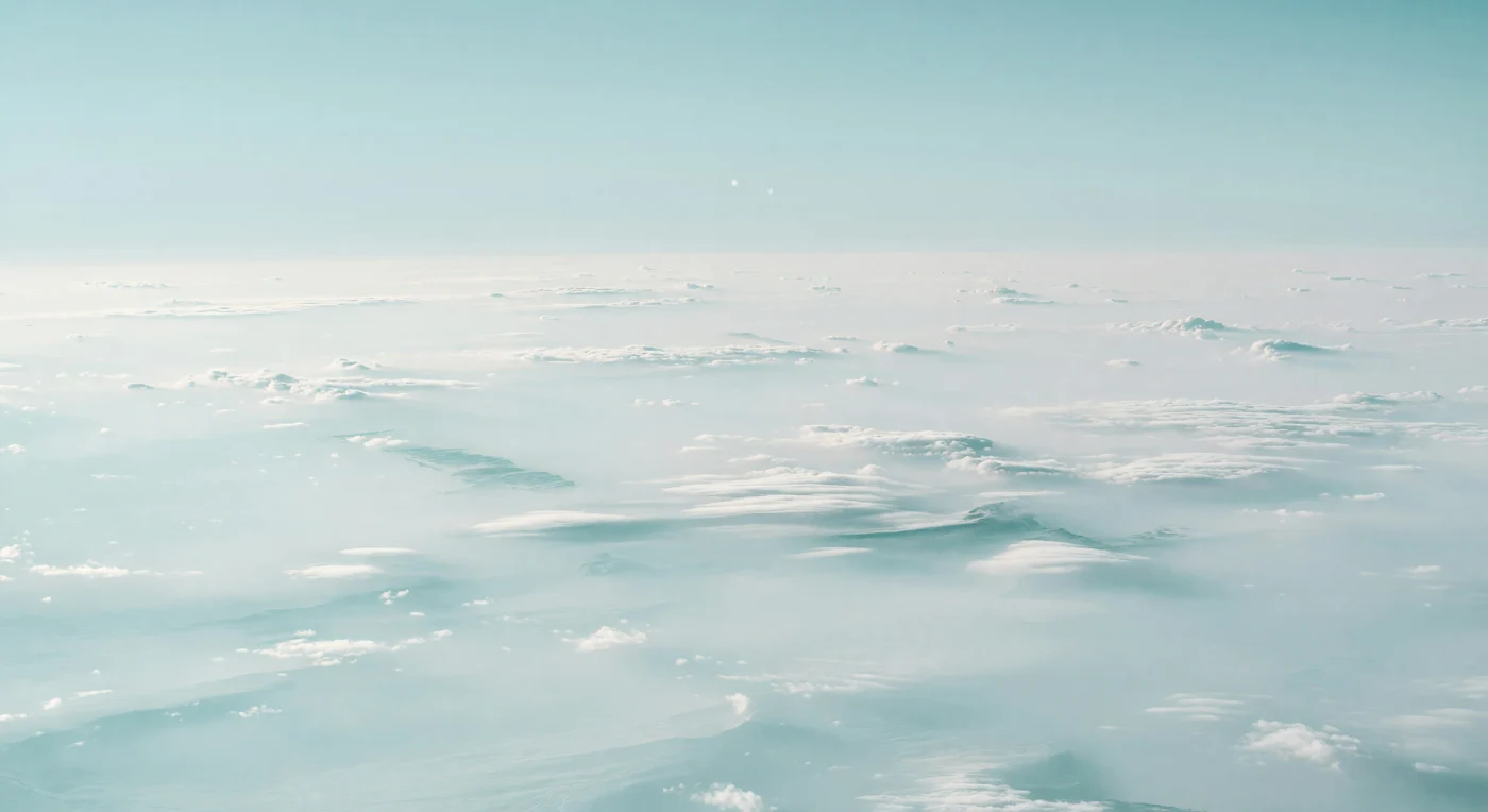 All around you, the world is nothing but atmosphere: a vast, pale white-cyan expanse of softly layered haze and faintly mottled cloud tops, their shallow swells and scalloped turbulence fading into a horizon so fog-bright it disappears into the sky. The scene is lit by an extraordinarily distant Sun, leaving the hydrogen-helium air and methane-rich aerosols washed in cold turquoise and silver-blue tones with almost no shadows, as if the entire landscape glows from within. These bright polar cloud layers are part of a high-latitude hood of condensed particles and methane haze suspended in the upper troposphere, where red light is absorbed and the visible world becomes desaturated, luminous, and eerily uniform. With no solid ground anywhere below—only deeper and denser atmospheric layers descending into high-pressure fluids—the immense, repeating fog banks and subdued cloud cells create a disorienting sense of standing inside an endless, frozen sea of sky.